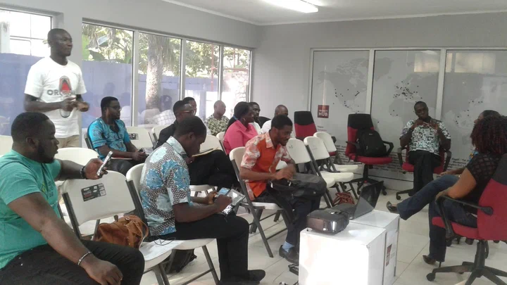 Young people seated in a meeting room listen as one man stands to speak